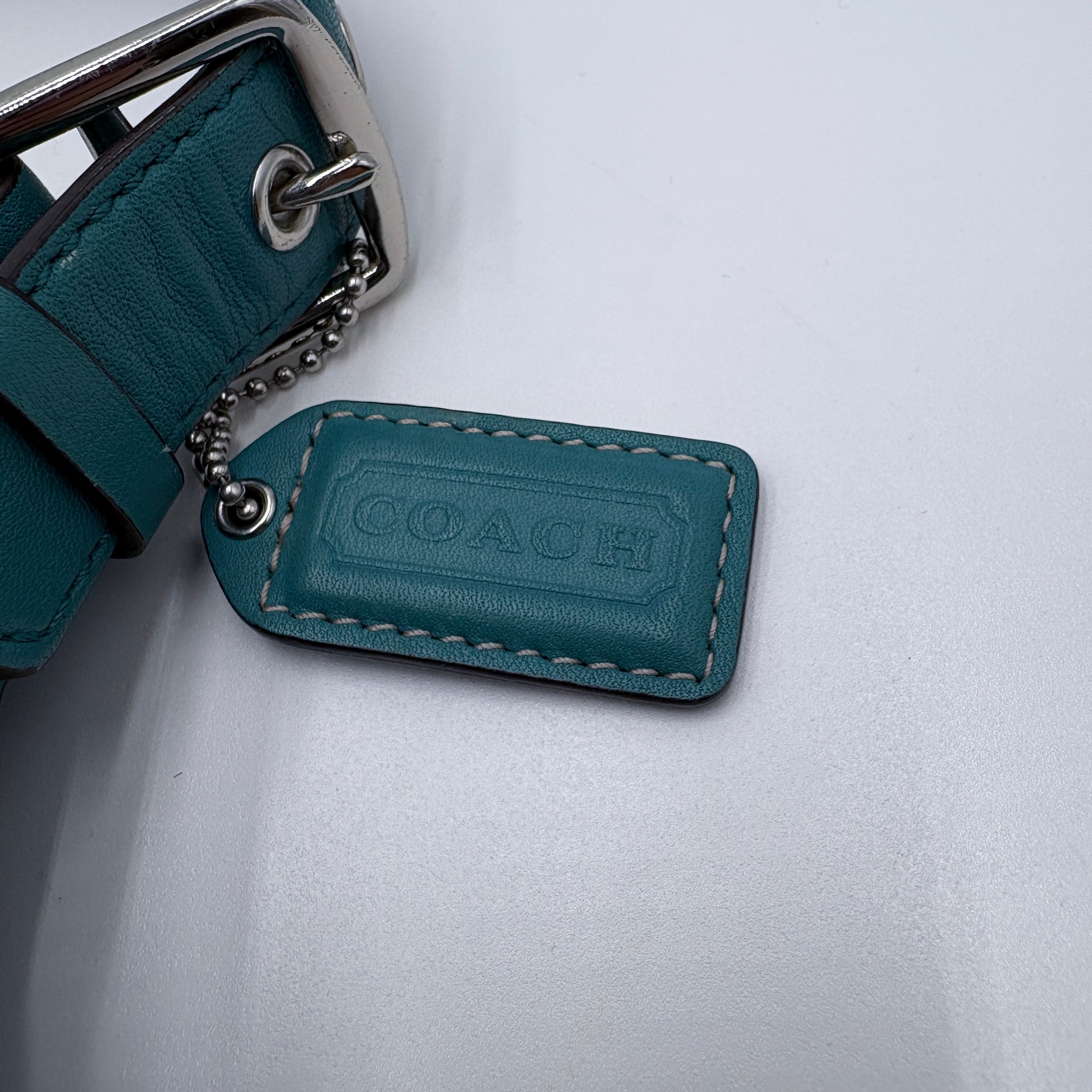 Teal leather strap with metal buckle and Coach logo tag on a light gray background