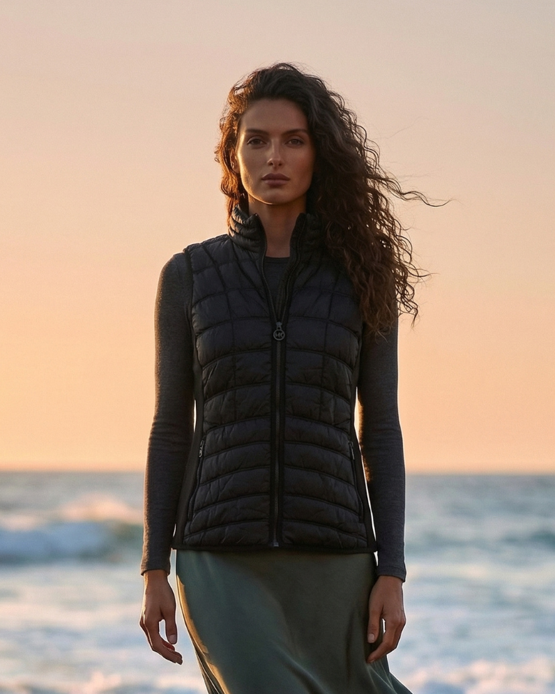 Woman wearing a black puffer vest on a beach at sunset with 'UrbanRevivalFashion.com' branding.