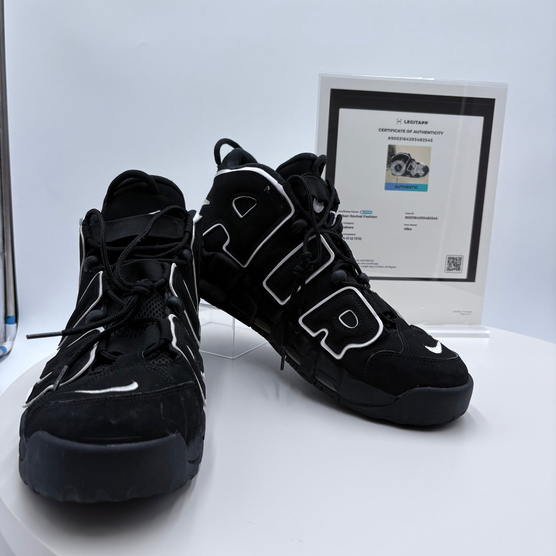 Black high-top sneakers with white accents on a white platform, accompanied by a certificate of authenticity.